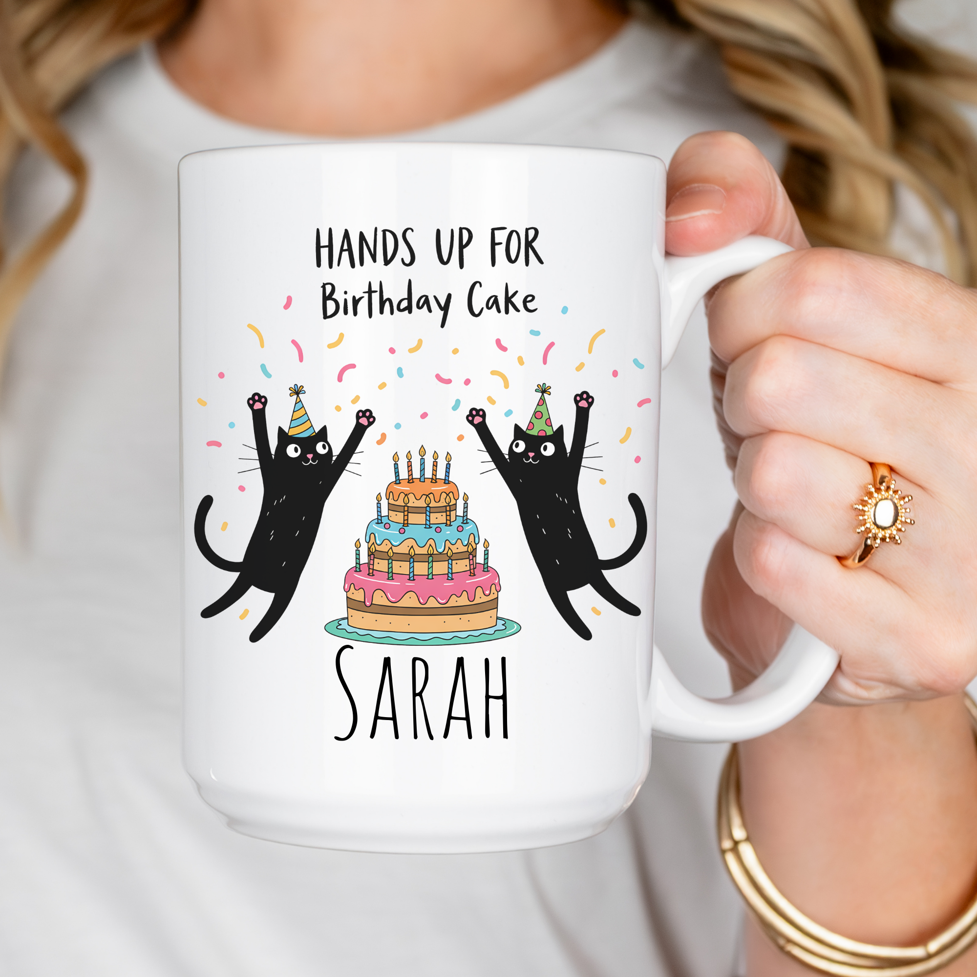 Person holding a 15oz. ceramic mug with an hand drawn design of two black cats wearing party hats high fiving over a birthday cake. Text 'Hands Up For Birthday Cake". Personalized with a name.