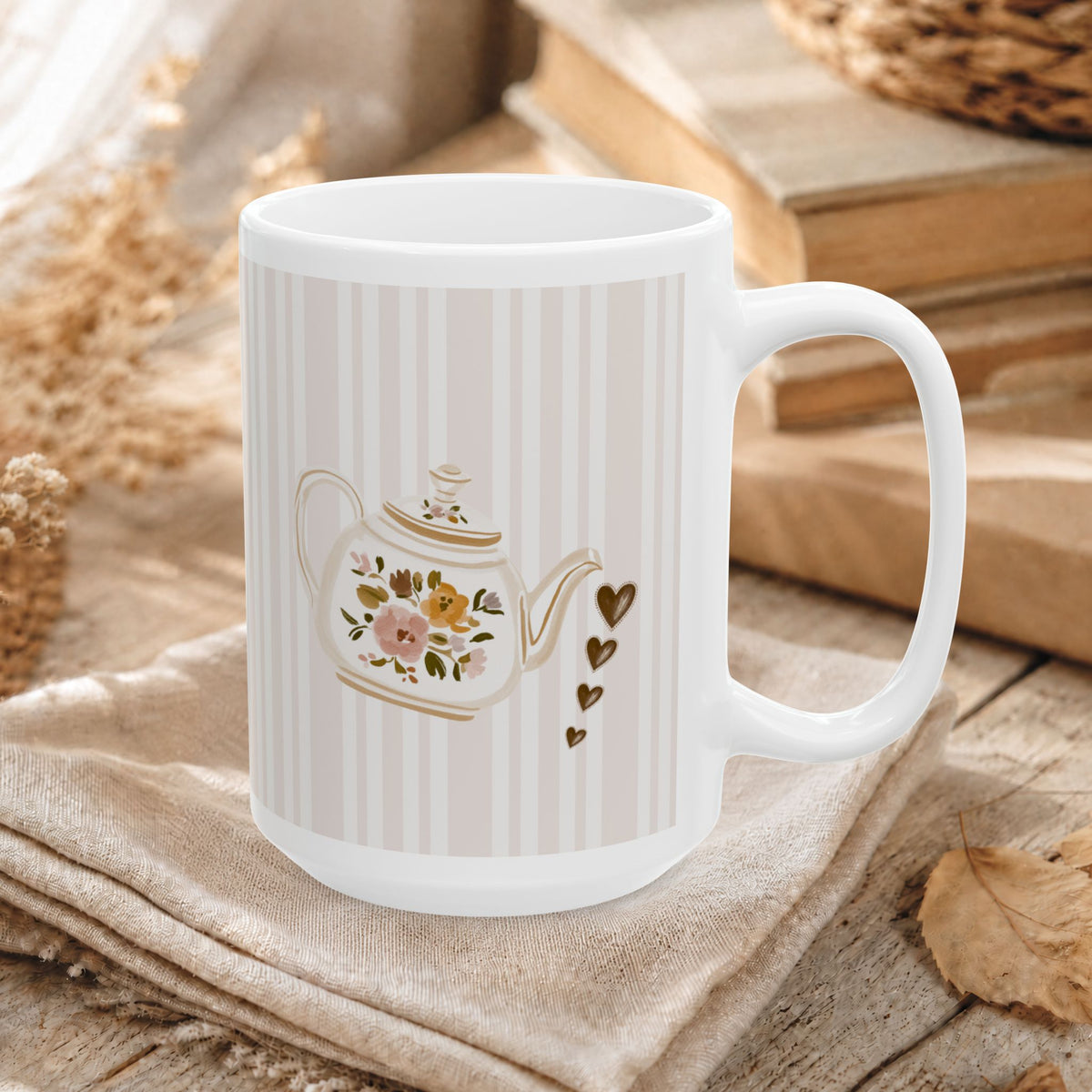 A white ceramic mug with a handle, decorated with a vintage-style floral teapot illustration and small gold hearts, resting on a beige cloth over a rustic wooden surface with books and dried flowers in the background.