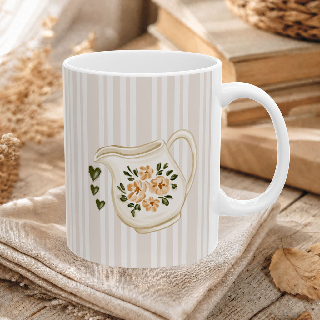 A white ceramic mug with vertical cream stripes and a painted cream pitcher floral design, displayed on a beige cloth over a rustic wooden surface with books and dried leaves in the background.