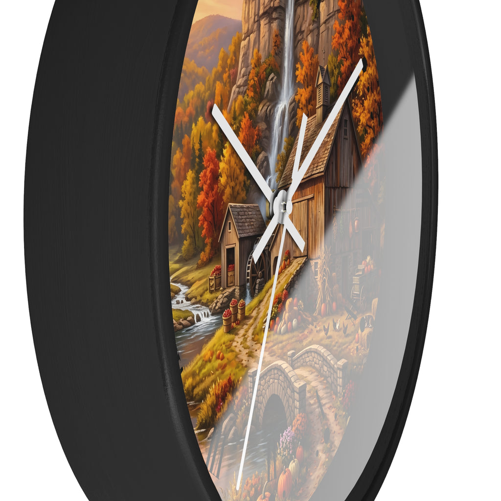 Round wall clock with a black frame and a scenic autumn landscape print featuring a rustic cabin, waterfall, stream, bridge, and colorful fall foliage. The clock is shown against a plain white background.