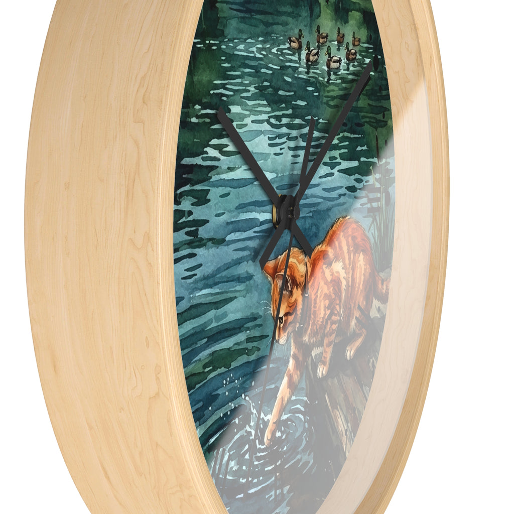 Oval wall clock with a light natural wood frame and a printed watercolor-style center depicting a orange cat in blue-green water with ducks overhead, shown on a white background.