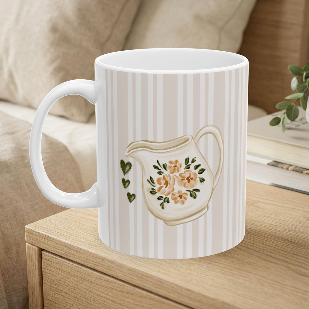 A white ceramic mug with vertical cream stripes and a printed illustration of a cream-colored teapot with peach flowers and green leaves, shown sitting on a wooden bedside table in a cozy bedroom setting.
