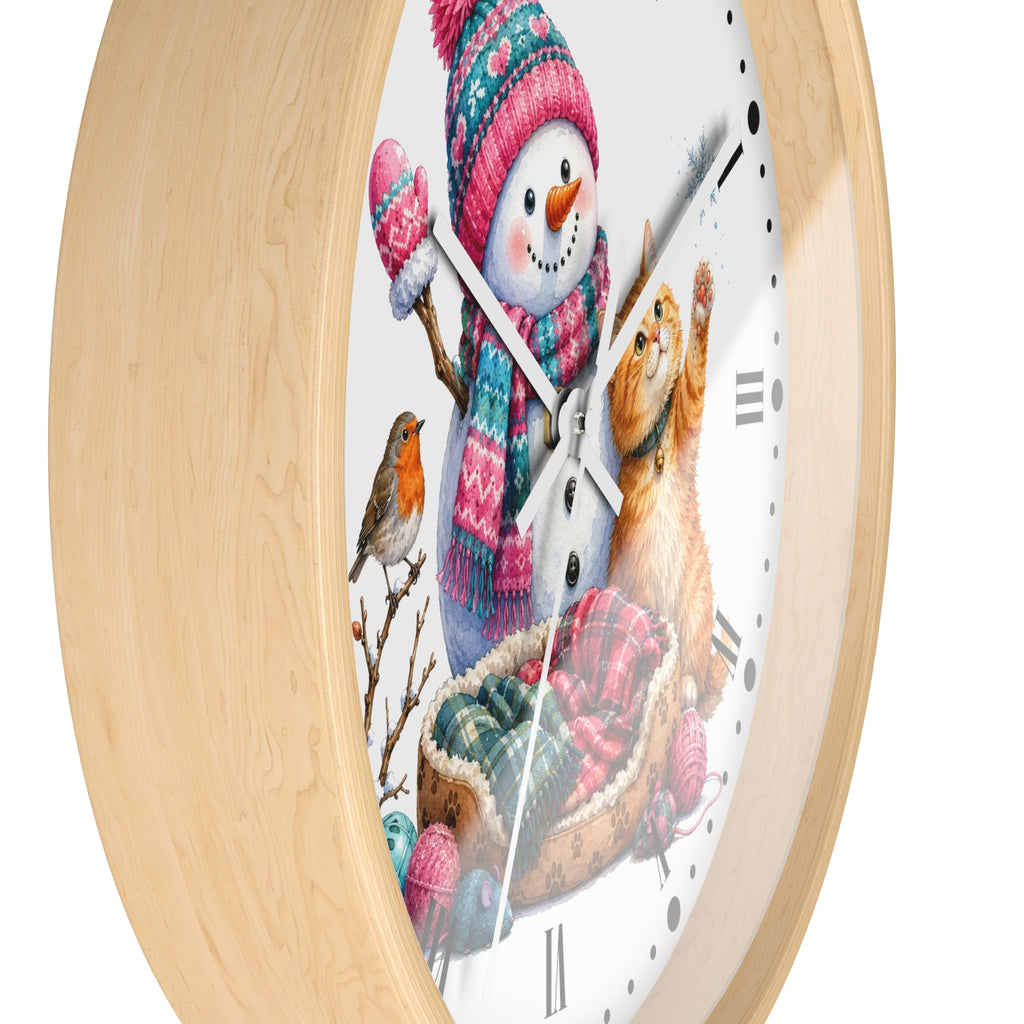 A round wall clock with a light natural wood frame and a white face featuring a winter illustration of a snowman, a ginger cat in a sled, a small bird, and colorful knit accessories.
