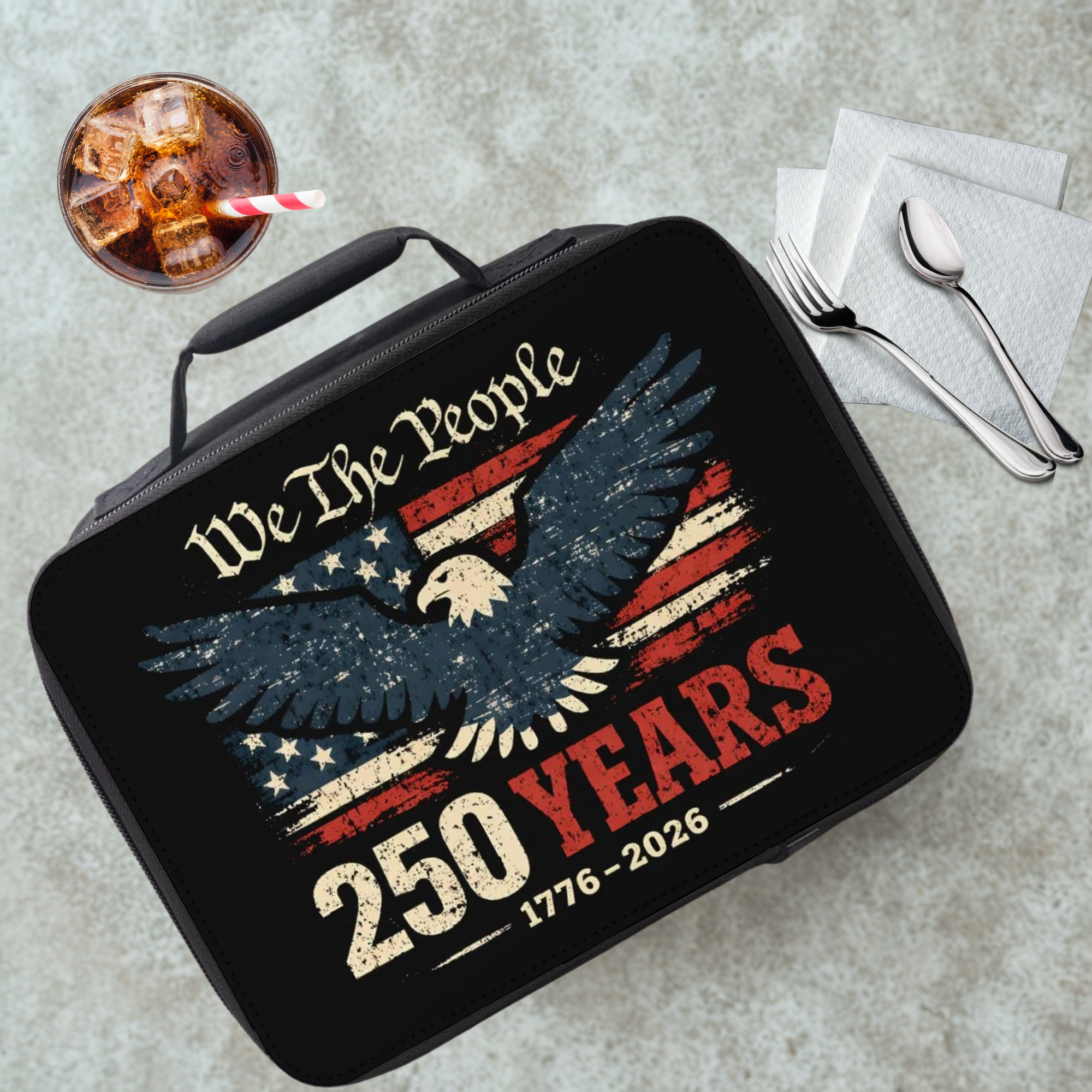 Black lunch box with 'We The People' and eagle design on a table with a drink and utensils.