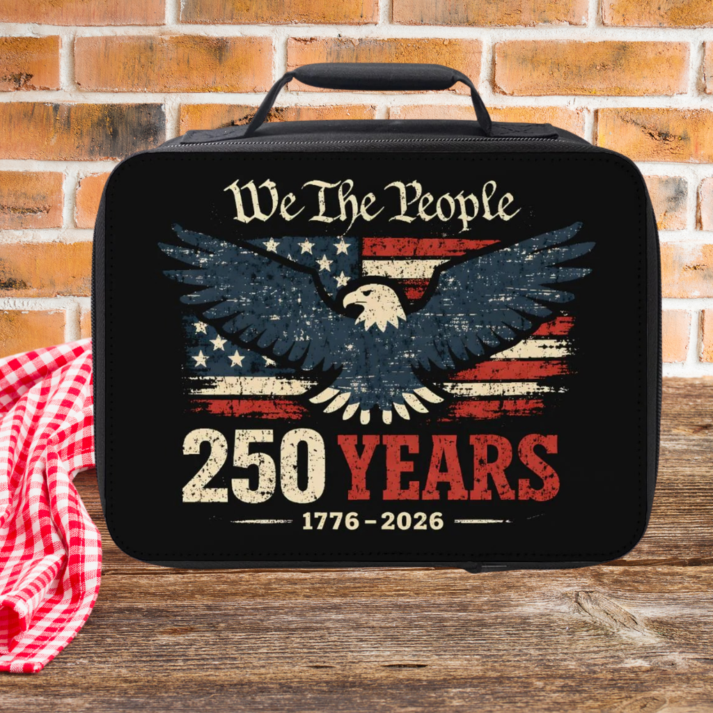 Black lunch bag with eagle design and 'We The People 250 Years' text on a wooden picnic table against a brick wall background