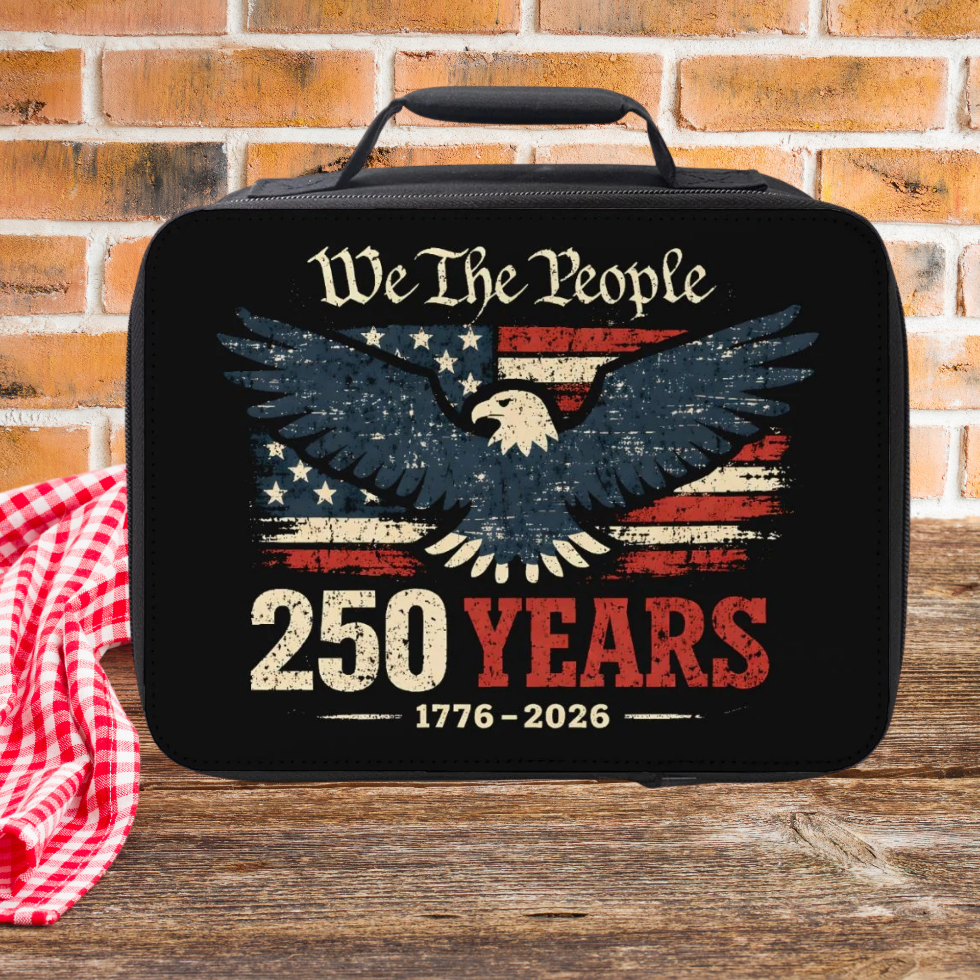 Black lunch bag with eagle design and 'We The People 250 Years' text on a wooden picnic table against a brick wall background
