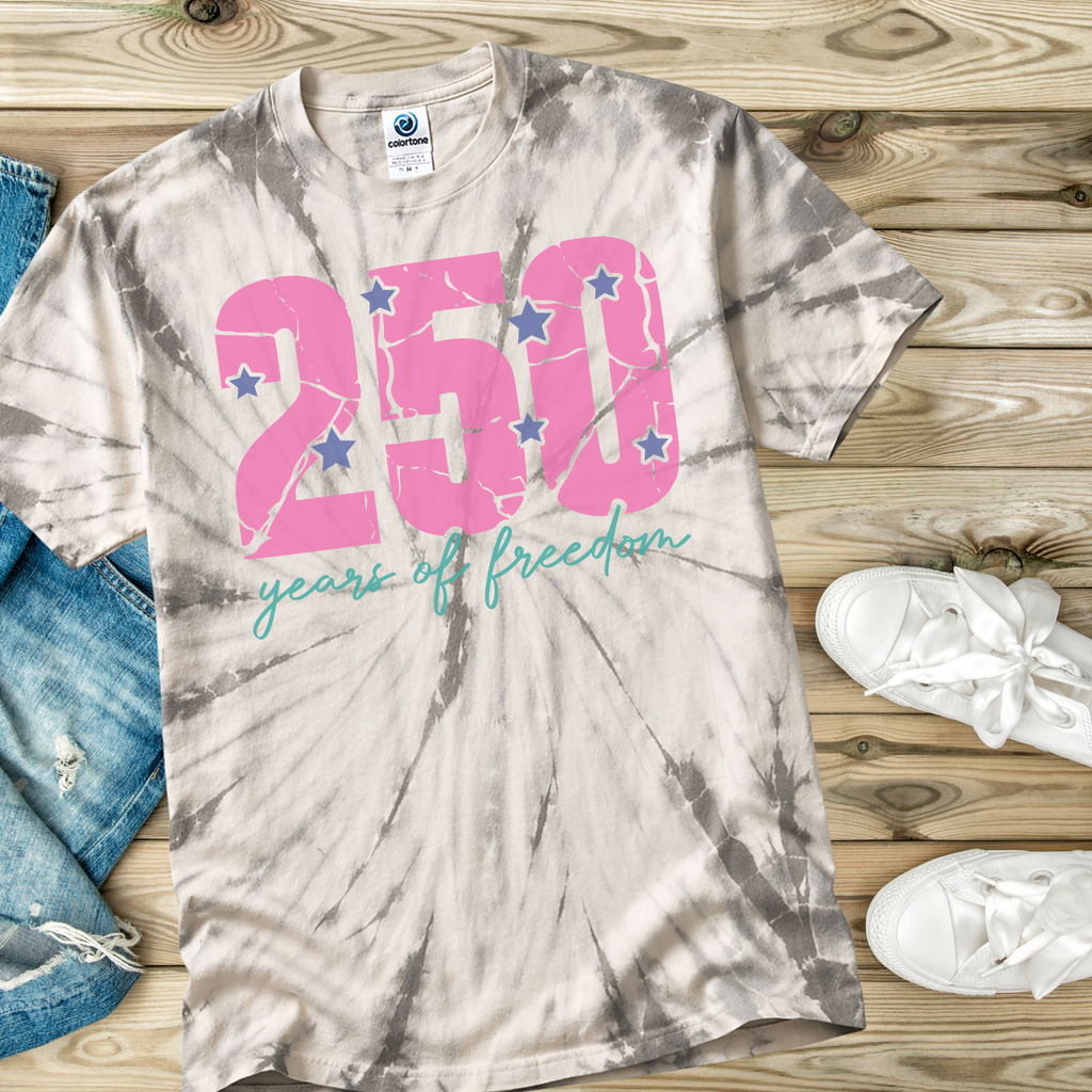 Tie-dye t-shirt with '250 years of freedom' text on a wooden surface with jeans and shoes.