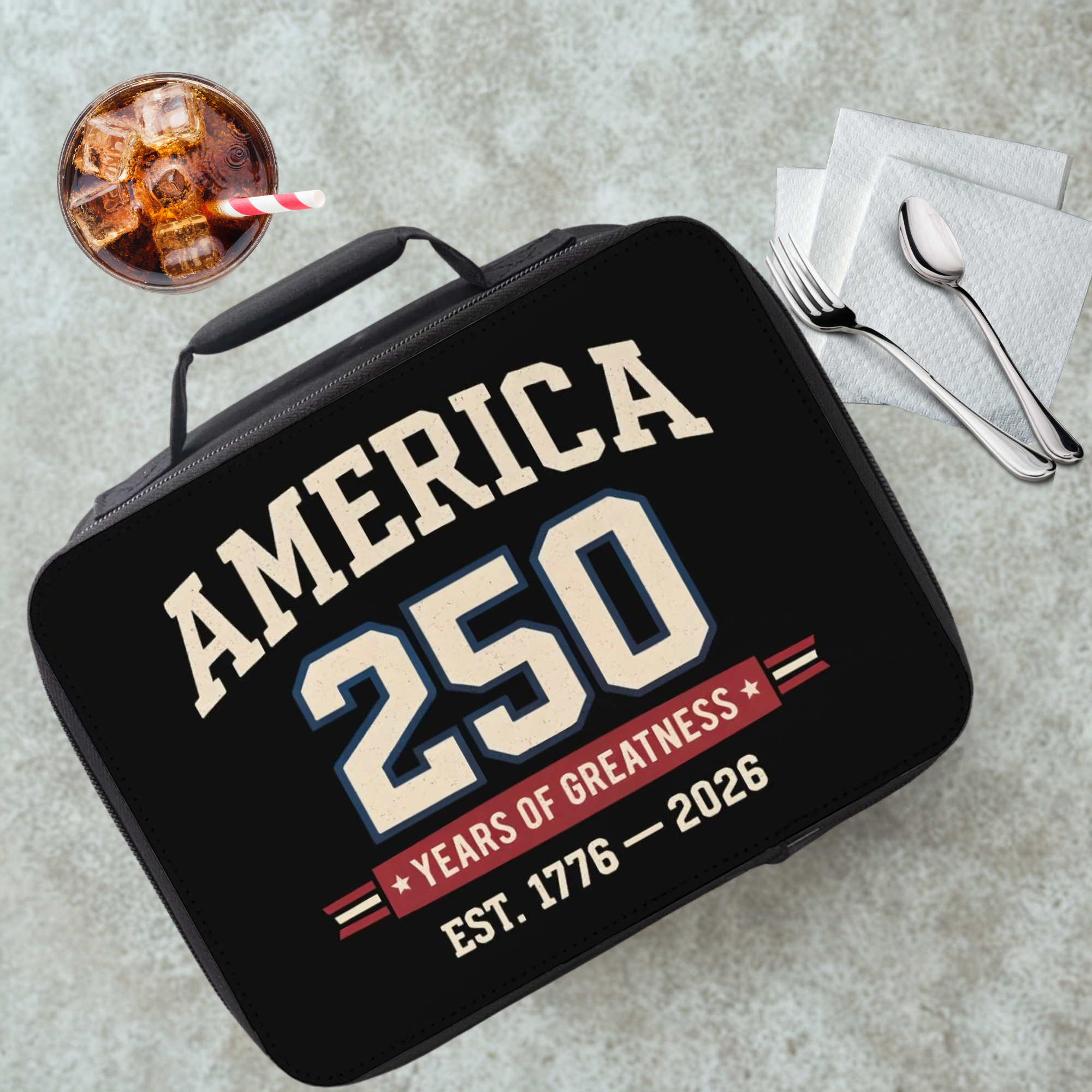 Black lunch box with 'America 250 Years of Greatness' text on a cafeteria table with a drink and utensils.
