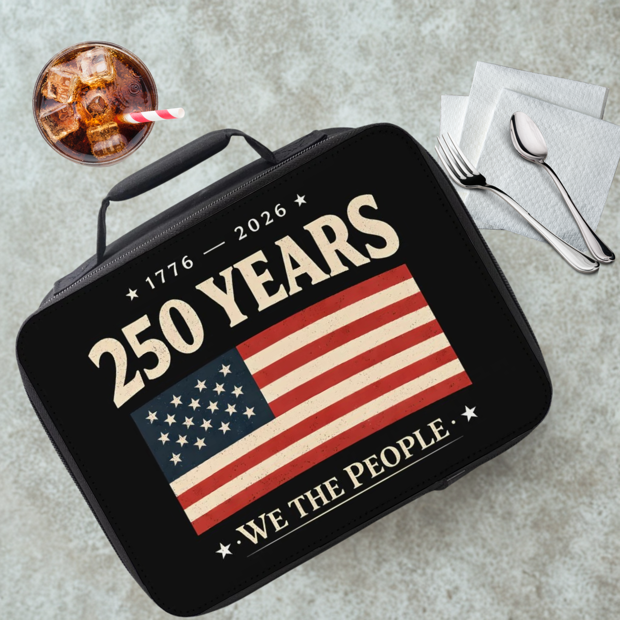 Black lunch box with American flag design and text on a grey table with a drink and utensils.
