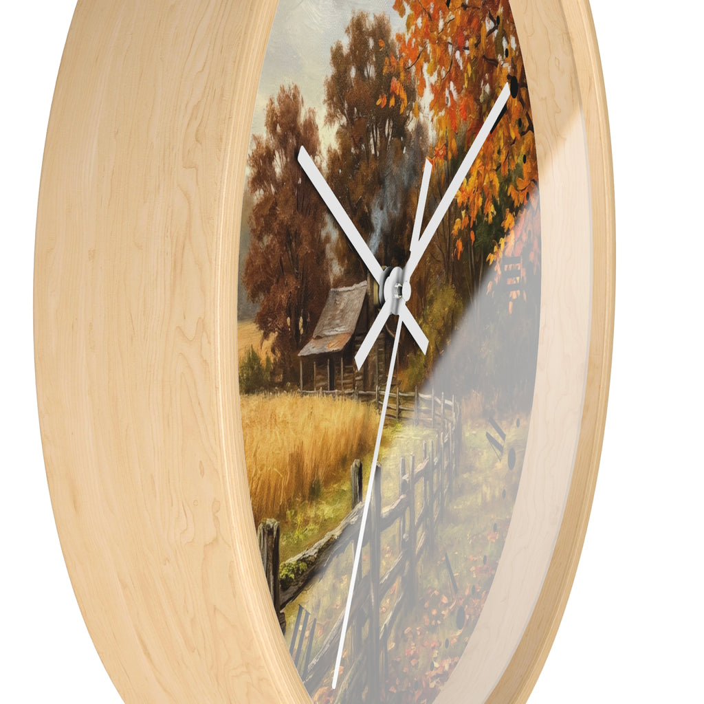 Round wall clock with a light natural wood frame and a printed autumn landscape scene featuring trees, a fence, a small cabin, and a field. The clock has white hands and is shown against a plain white background.