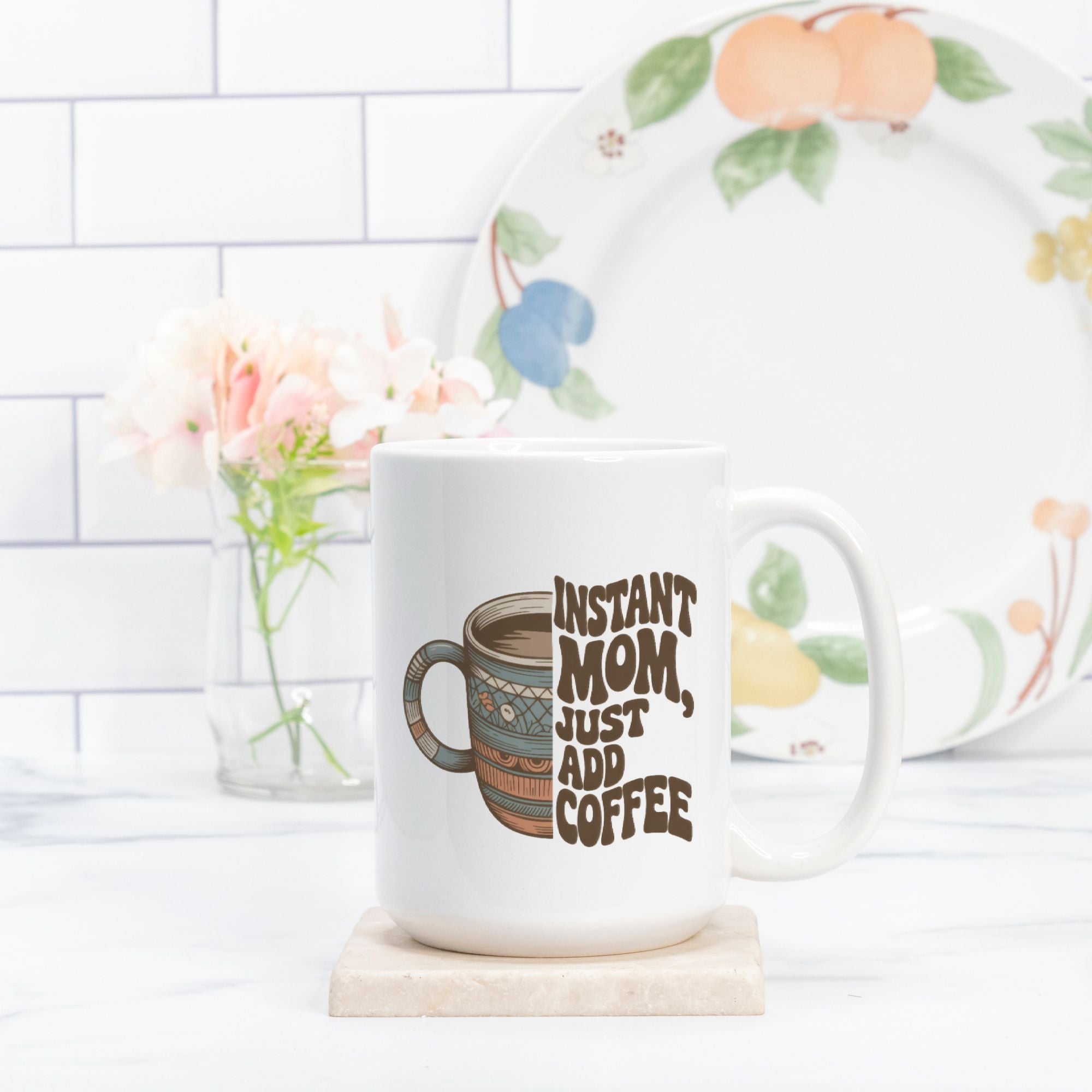 White mug with 'Instant Mom, Just Add Coffee' text on a coaster with a floral plate in the background.