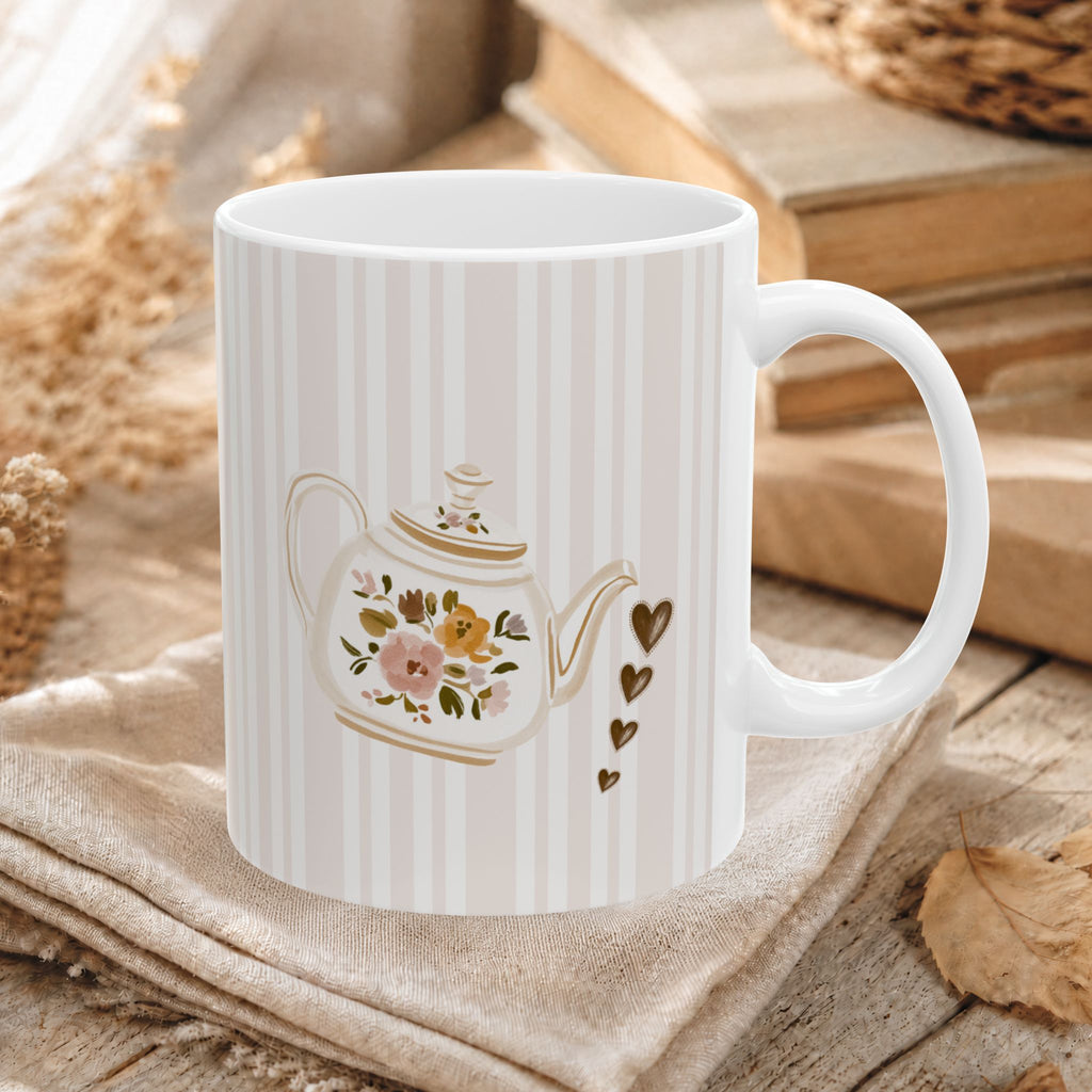 A white ceramic mug with vertical cream stripes and a printed teapot design with flowers, gold accents, and small hearts, styled on a beige cloth over a rustic wooden surface.