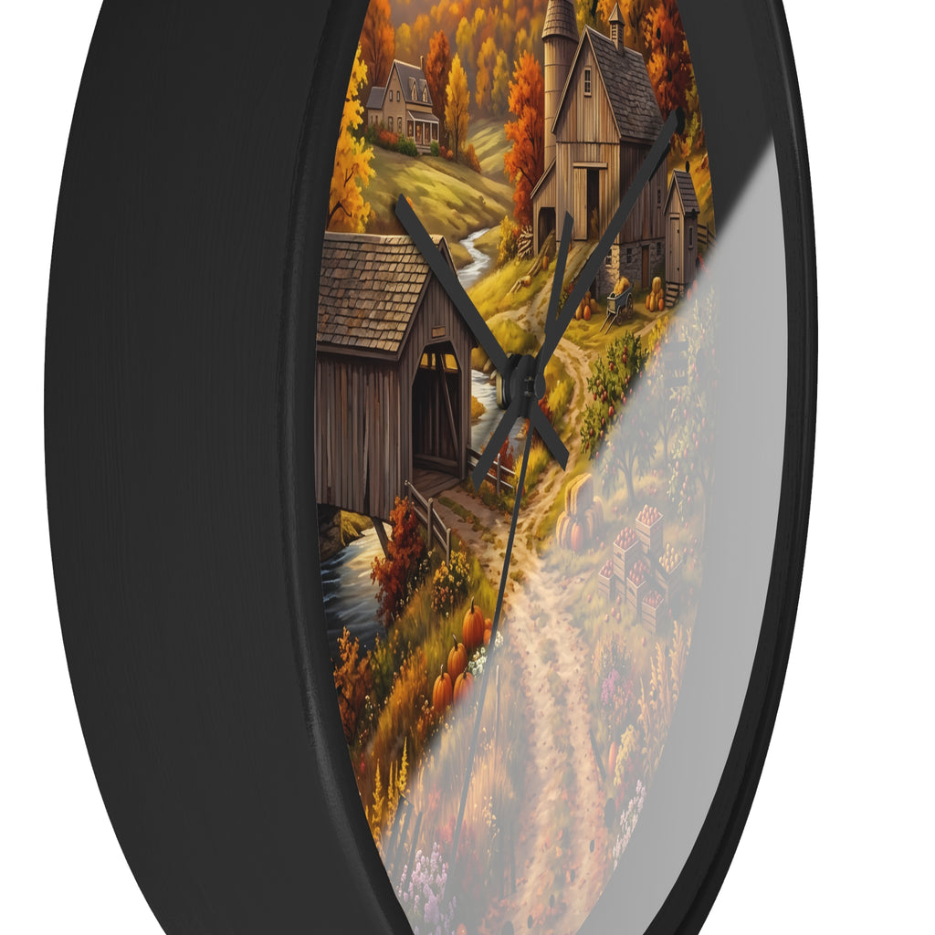 A round wall clock with a black frame and a printed autumn village scene on the face, featuring wooden houses, orange trees, a stream, and black clock hands against a white background.