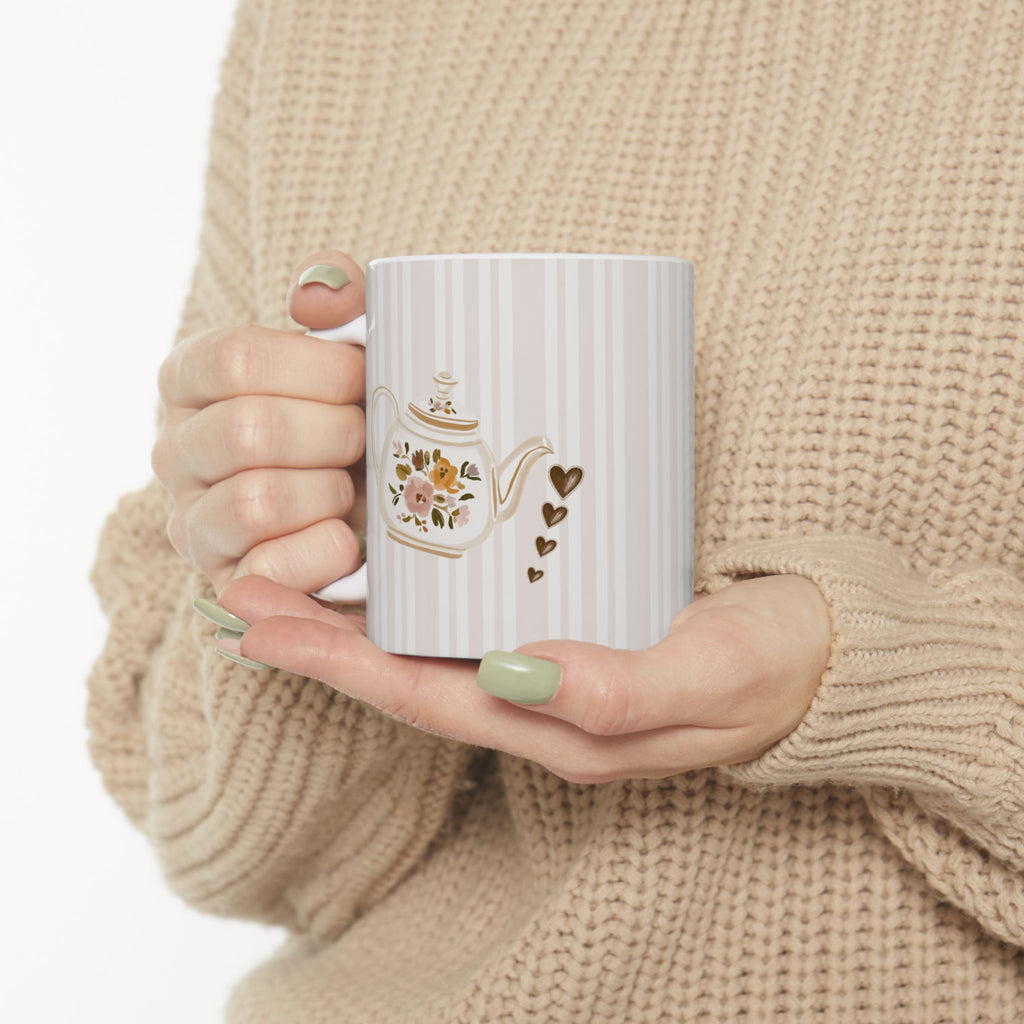 A person in a beige knit sweater holds a white mug with vertical stripes, a floral teapot illustration, and small gold hearts.