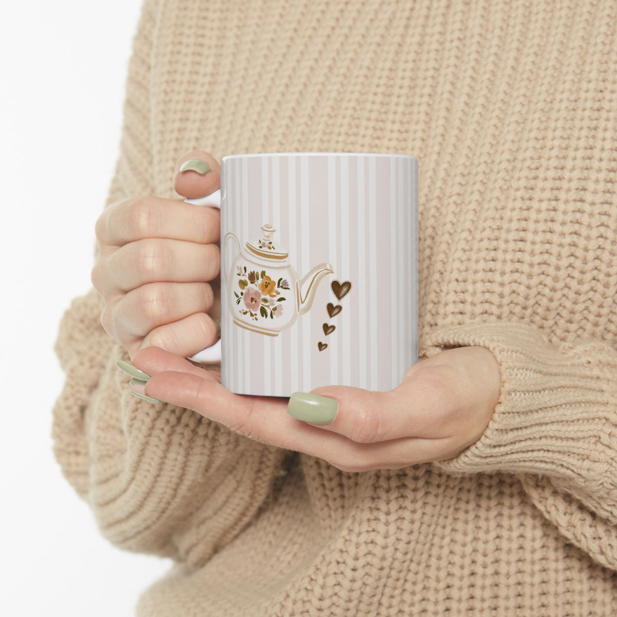 A person in a beige knit sweater holds a white mug with vertical stripes, a floral teapot illustration, and small gold hearts.