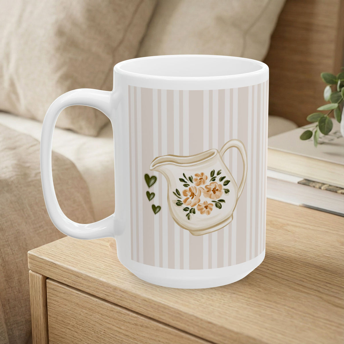 A white ceramic mug with a beige and white vertical stripe pattern, featuring a printed illustration of a cream-colored teapot with orange flowers and green leaves, alongside three small green hearts. The mug is shown sitting on a light wood table in a cozy living room setting.
