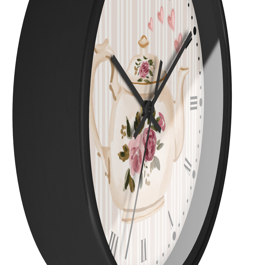 A close-up of a round wall clock with a black frame and a decorative dial featuring a cream teapot illustration with pink flowers and striped background.