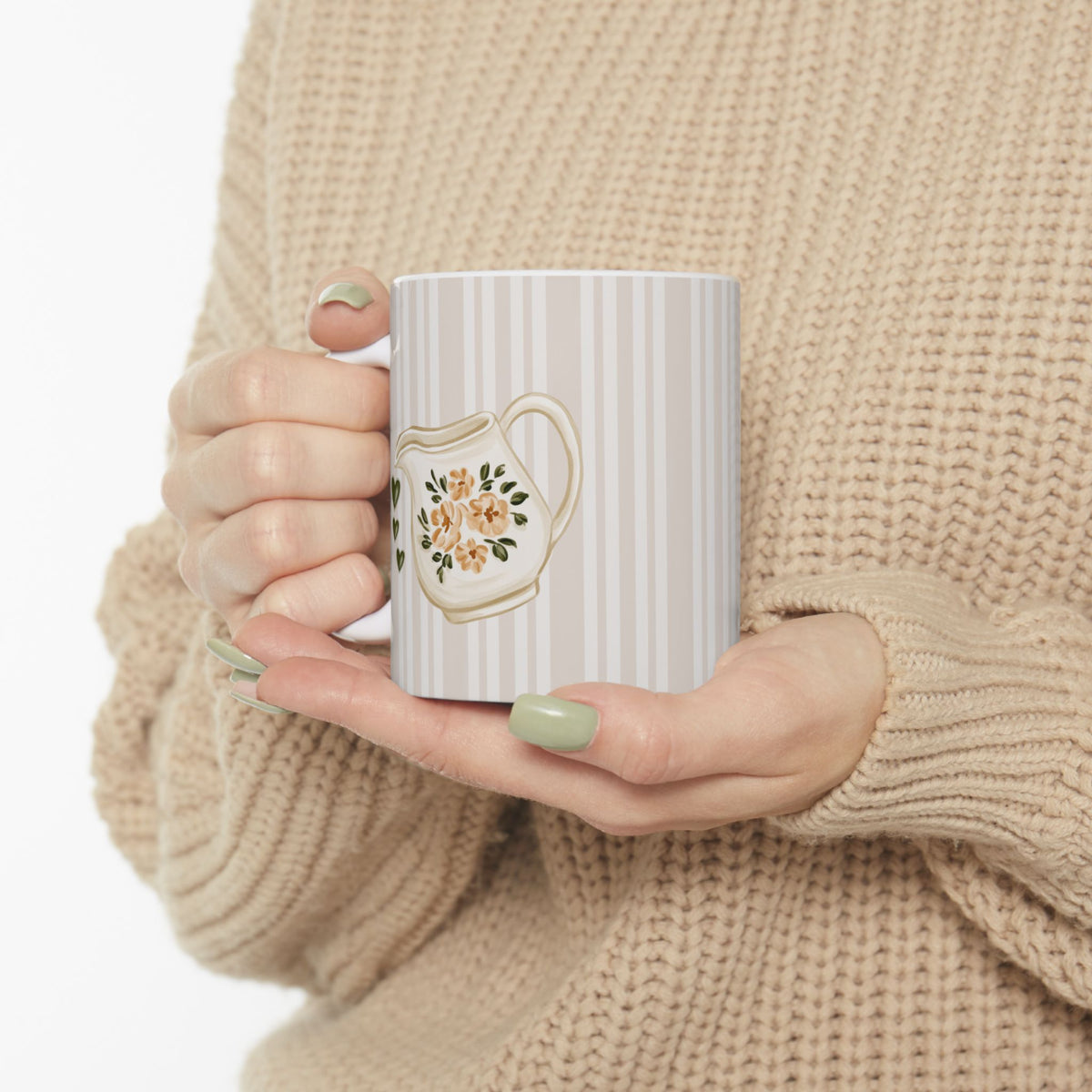 A person in a beige knit sweater holds a white ceramic mug with vertical gray stripes and a floral teapot illustration on the side.