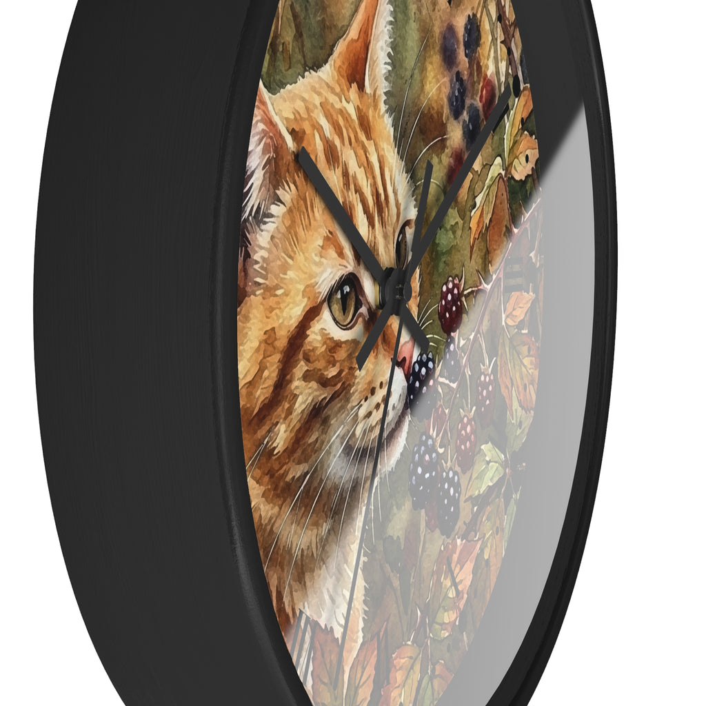 A round wall clock with a black frame and a printed design of an orange tabby cat surrounded by autumn leaves and blackberries on the face, shown against a plain white background.