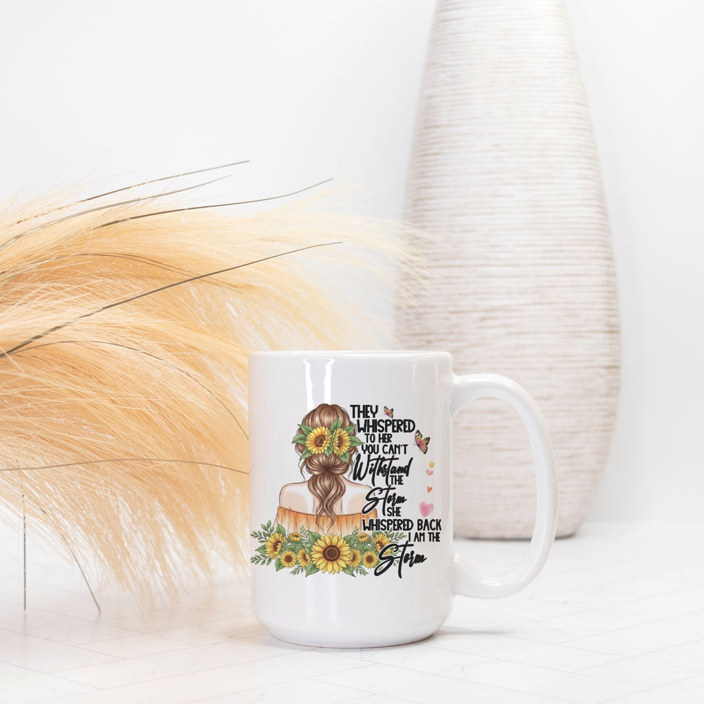 White mug with a design featuring sunflowers and text, placed on a white surface with a blurred background.