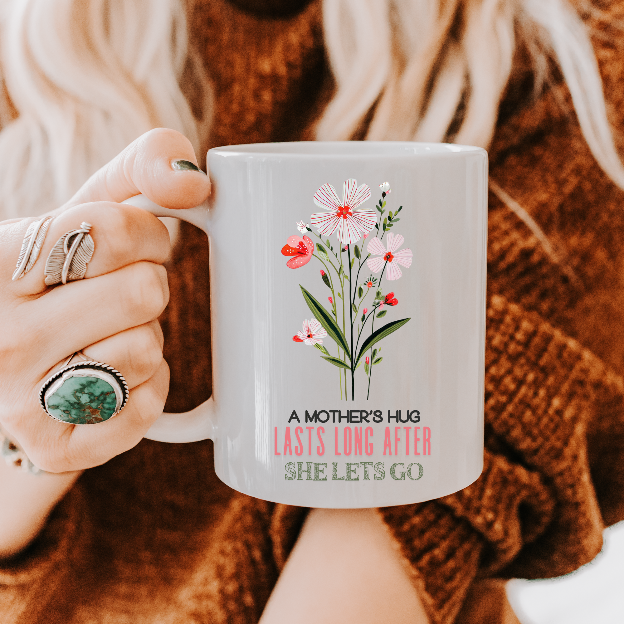 Person holding an 11oz mug with floral design and text 'A Mother's Hug Lasts Long After She Lets Go, and wearing a brown sweater.