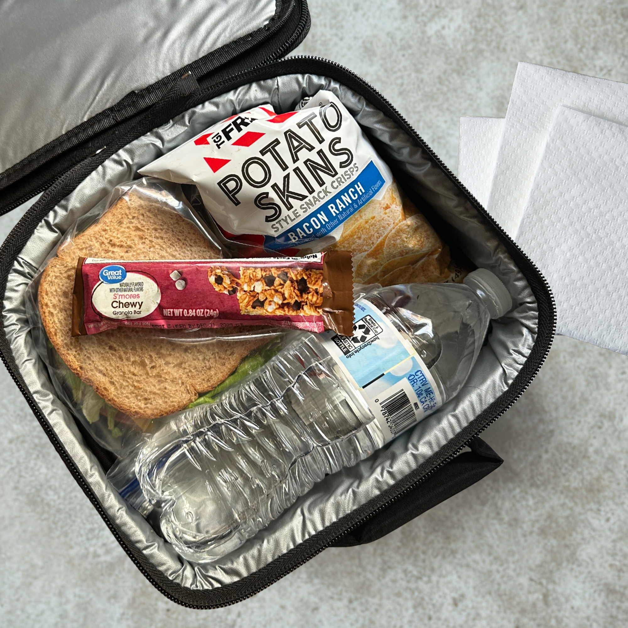 Lunchbox filled with sandwich, chips, and water on a cafeteria table with napkins.