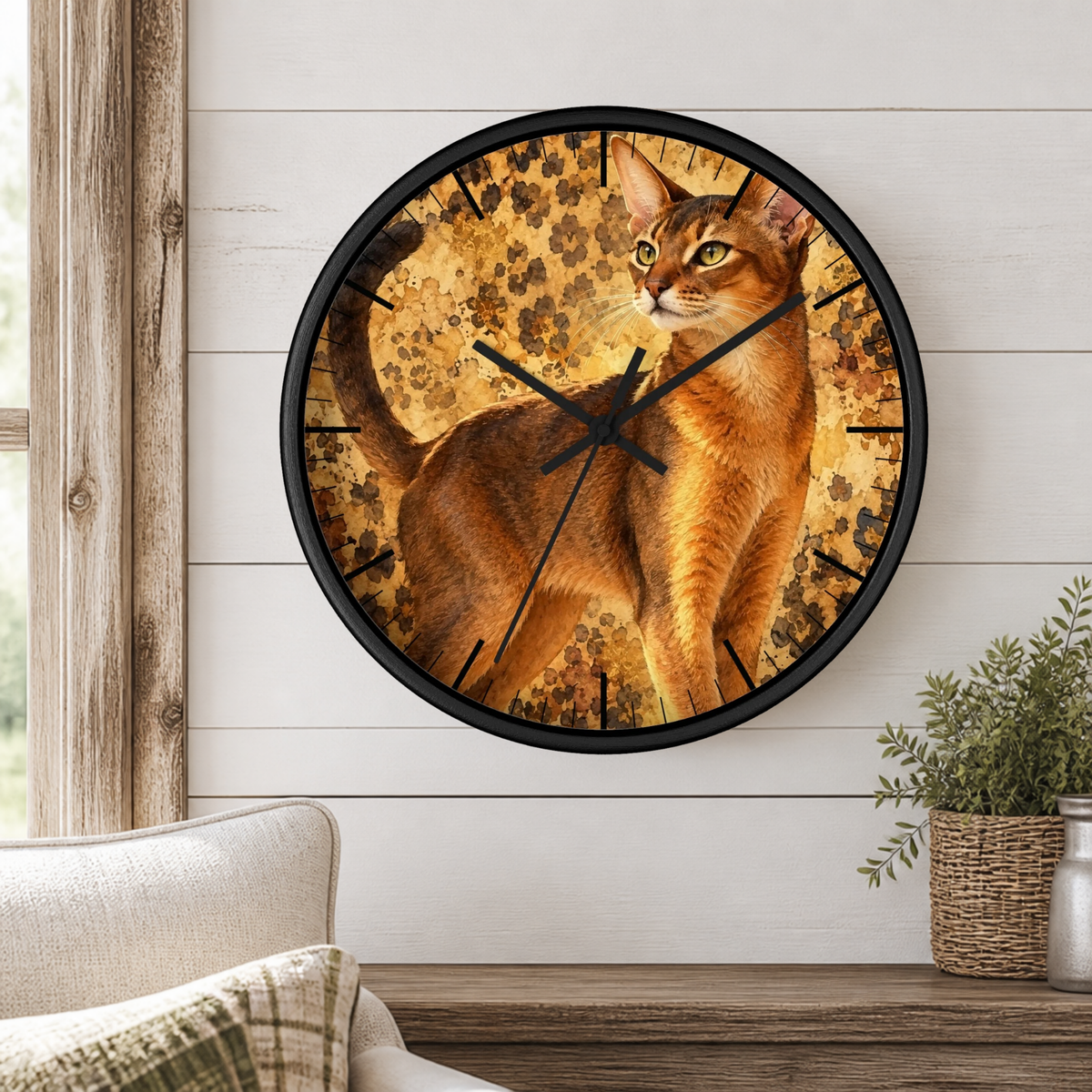 Round wall clock with a black frame and a printed cat face design, mounted on a white shiplap wall above a wooden shelf with a plant and part of a sofa visible.