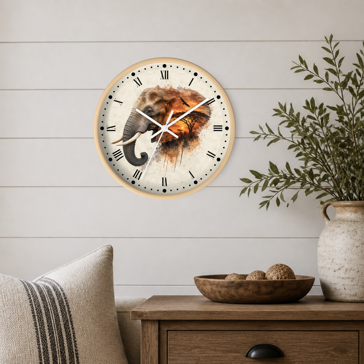 A round wall clock with a light wood frame and an elephant illustration on the face, mounted on a white shiplap wall above a wooden side table with a vase and bowl.