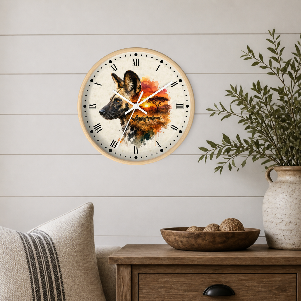 A round wall clock with a light wood frame hangs on a white shiplap wall above a wooden side table. The clock face features a painted African wild dog over an orange and yellow landscape, with black Roman numerals and white hands.