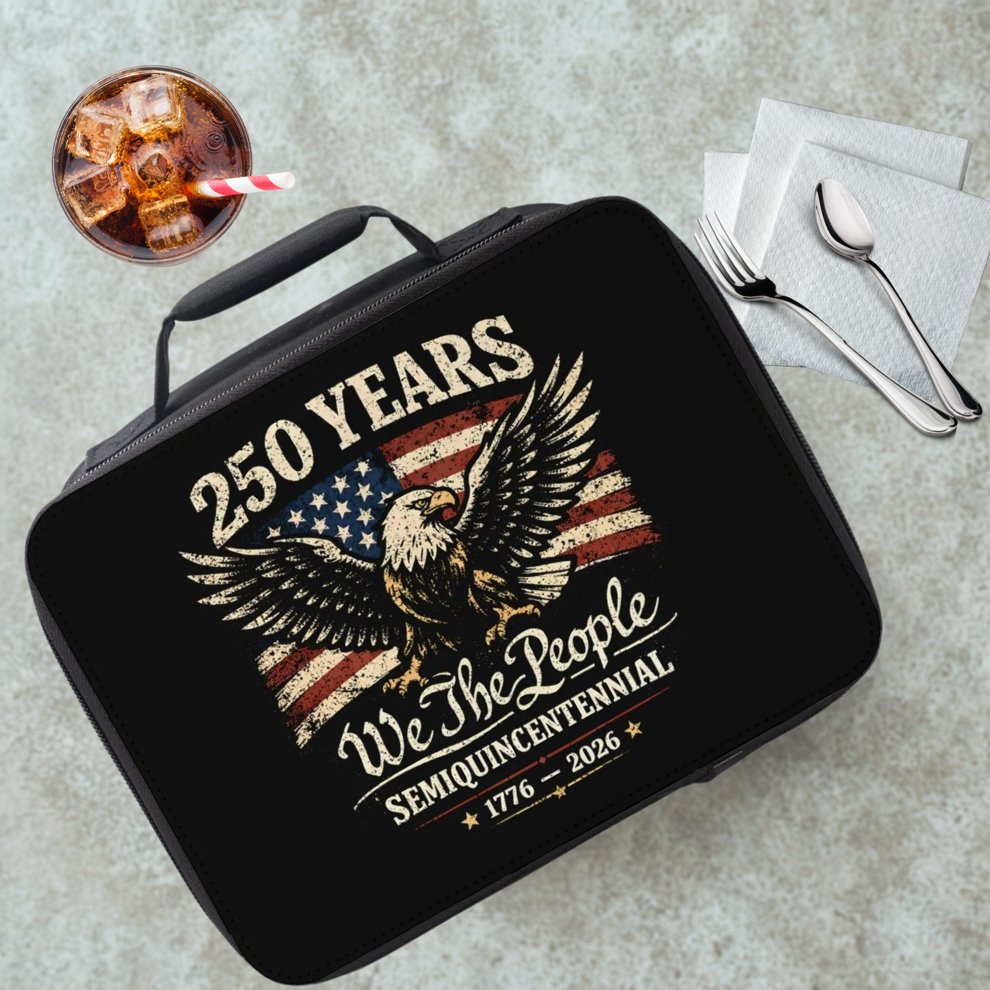 Black insulated lunch box with American flag and eagle design on a cafeteria table with a drink and utensils.