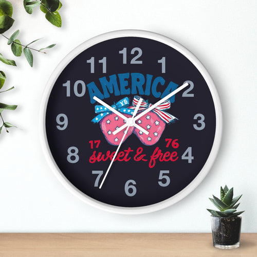 Round white wall clock with matching white hands and decorative strawberry fourth of July design and text on a white wall with plants.