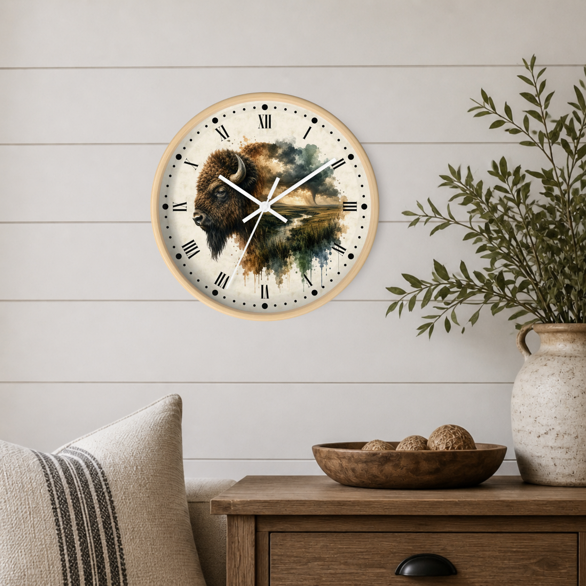A round wall clock with a watercolor bison design on the face, mounted on a white shiplap wall above a wooden side table with a plant and decorative bowl.