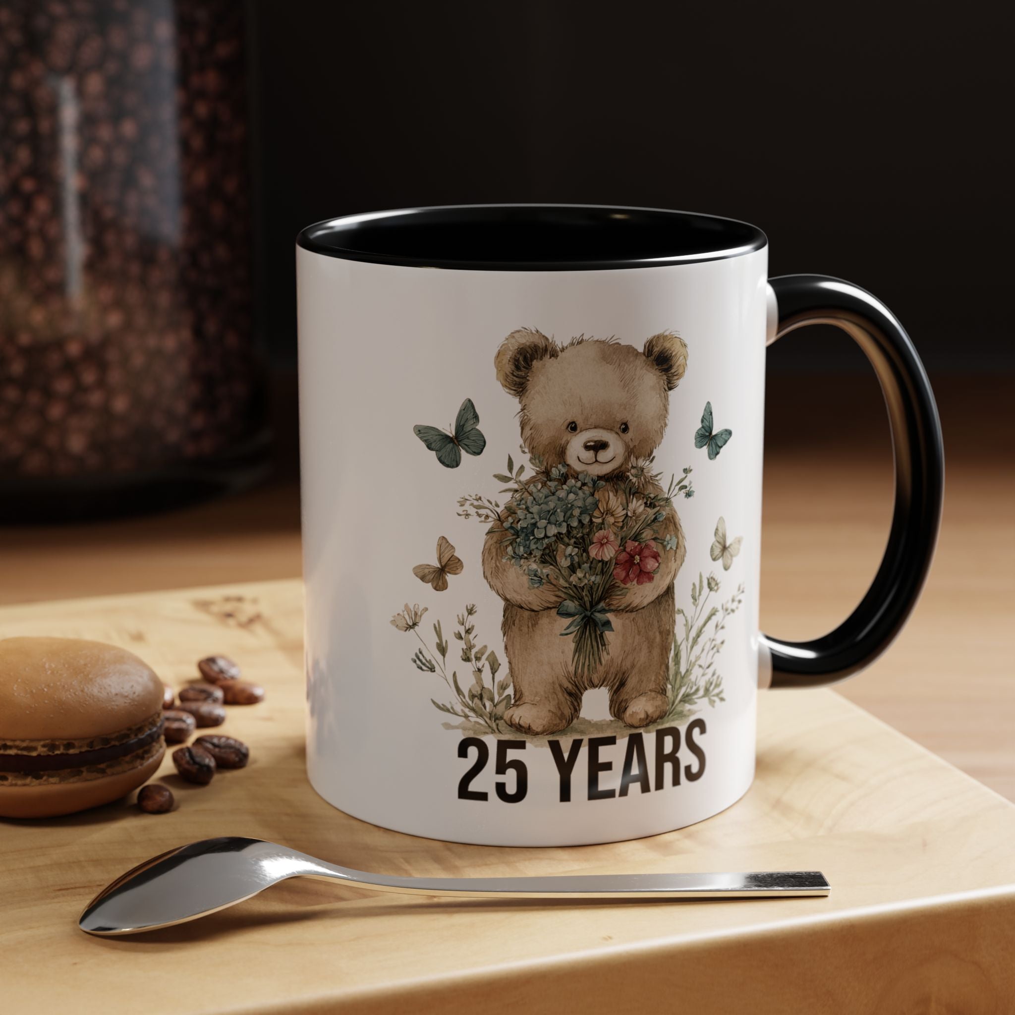 Mug with teddy bear design and '25 Years' text on a wooden surface with coffee beans and a spoon.
