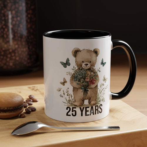 Mug with teddy bear design and '25 Years' text on a wooden surface with coffee beans and a spoon.