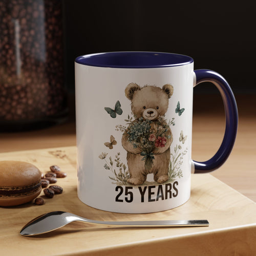 Mug with teddy bear design and '25 Years' text on a wooden surface with coffee beans and a spoon.