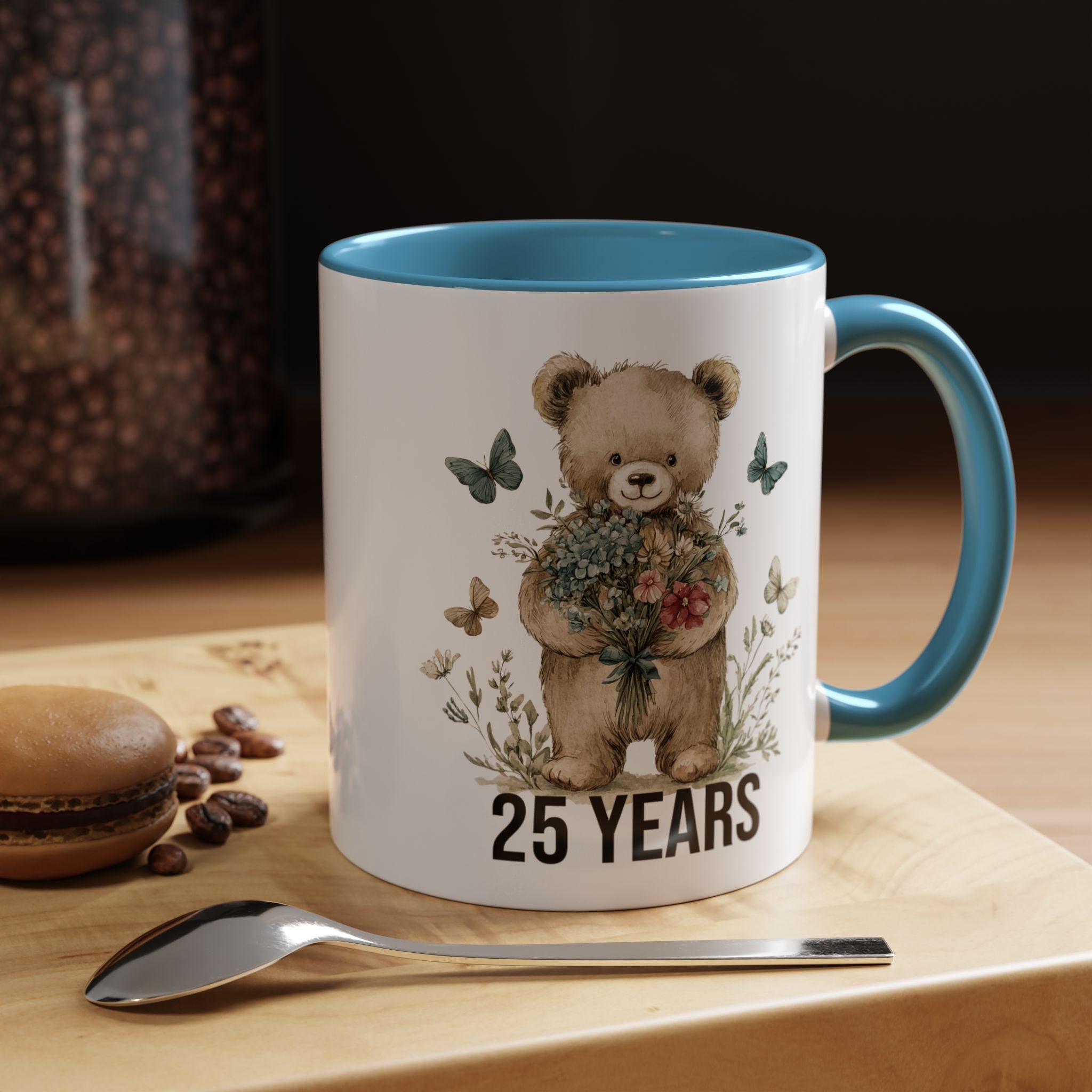Mug with teddy bear design and '25 Years' text on a wooden surface with coffee beans and a spoon.
