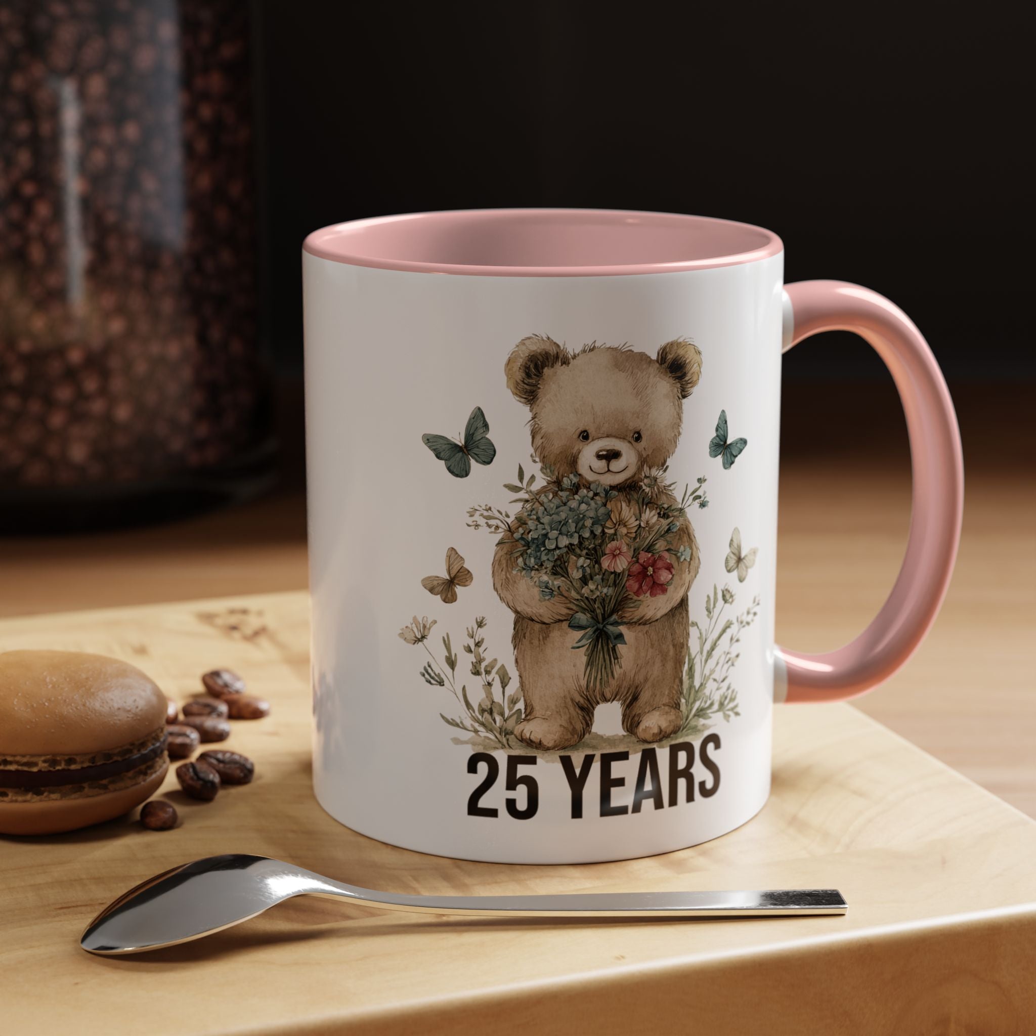Mug with teddy bear design and '25 Years' text on a wooden surface with coffee beans and a spoon.