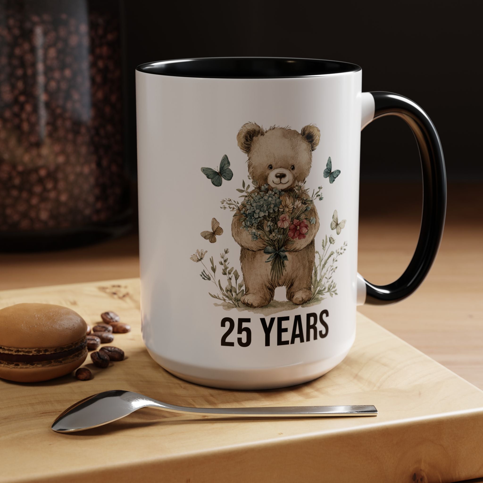 Mug with teddy bear design and '25 Years' text on a wooden surface with coffee beans and a macaron.