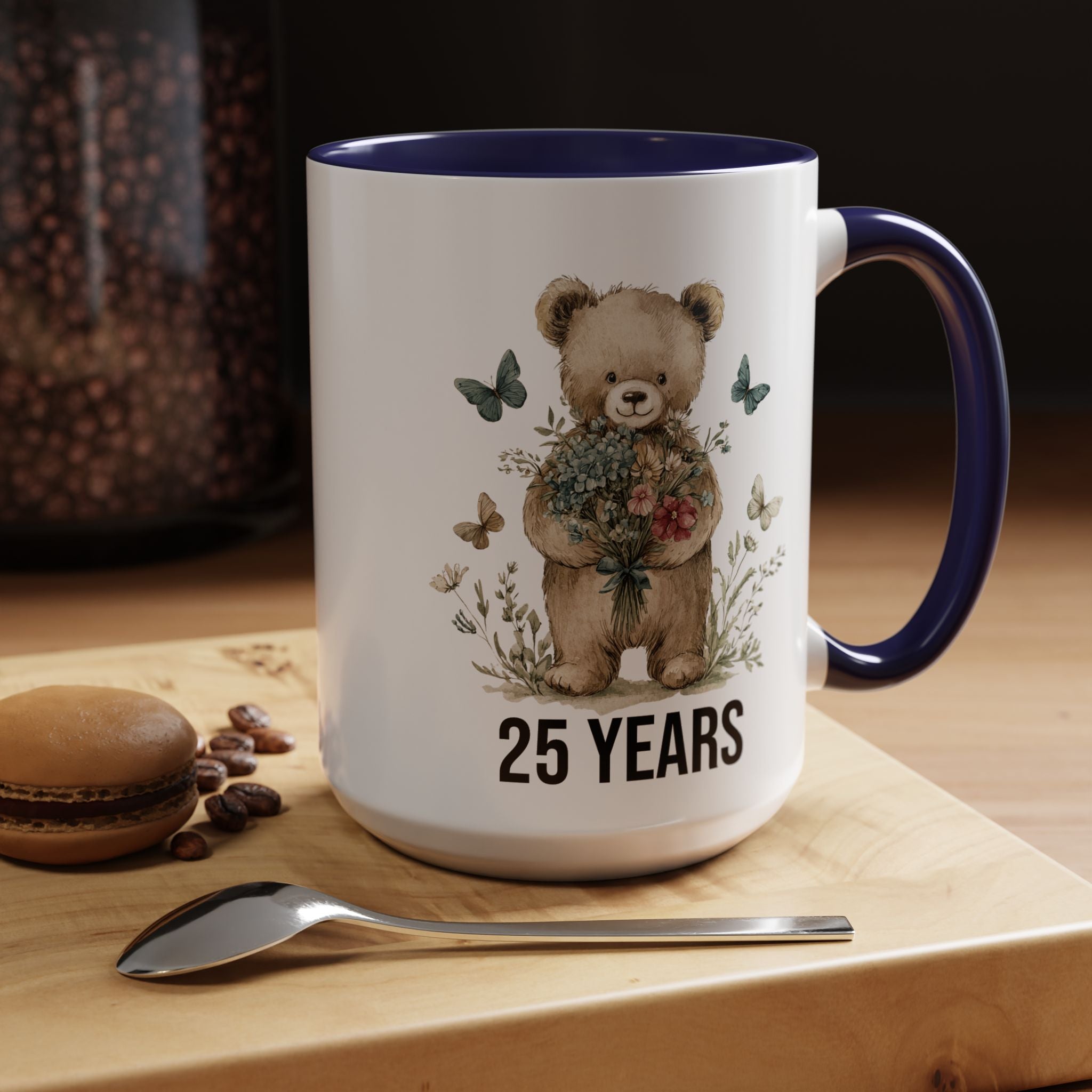 Mug with teddy bear design and '25 Years' text on a wooden surface with coffee beans and a spoon.