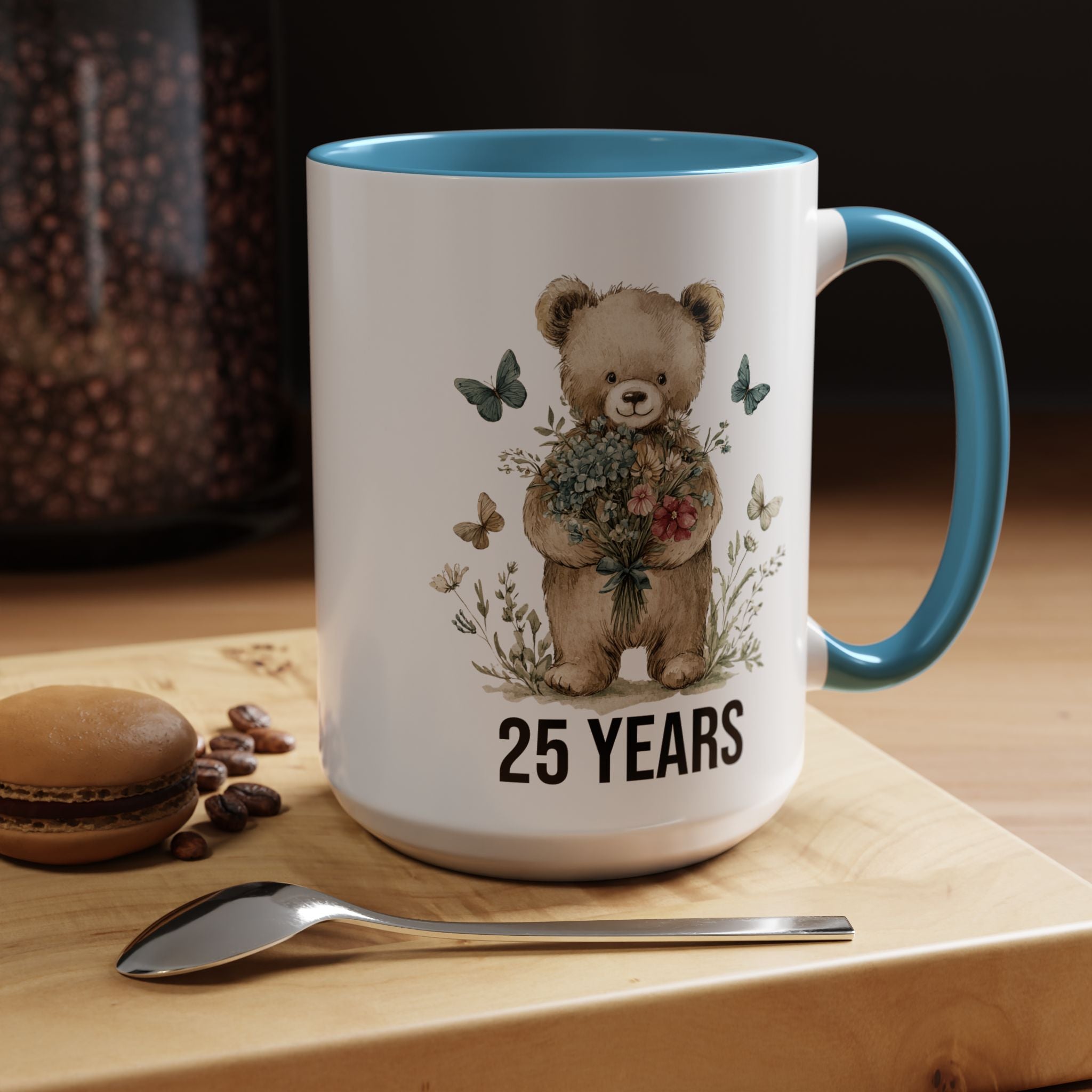 Mug with teddy bear design and '25 Years' text on a wooden surface with coffee beans and a macaron.