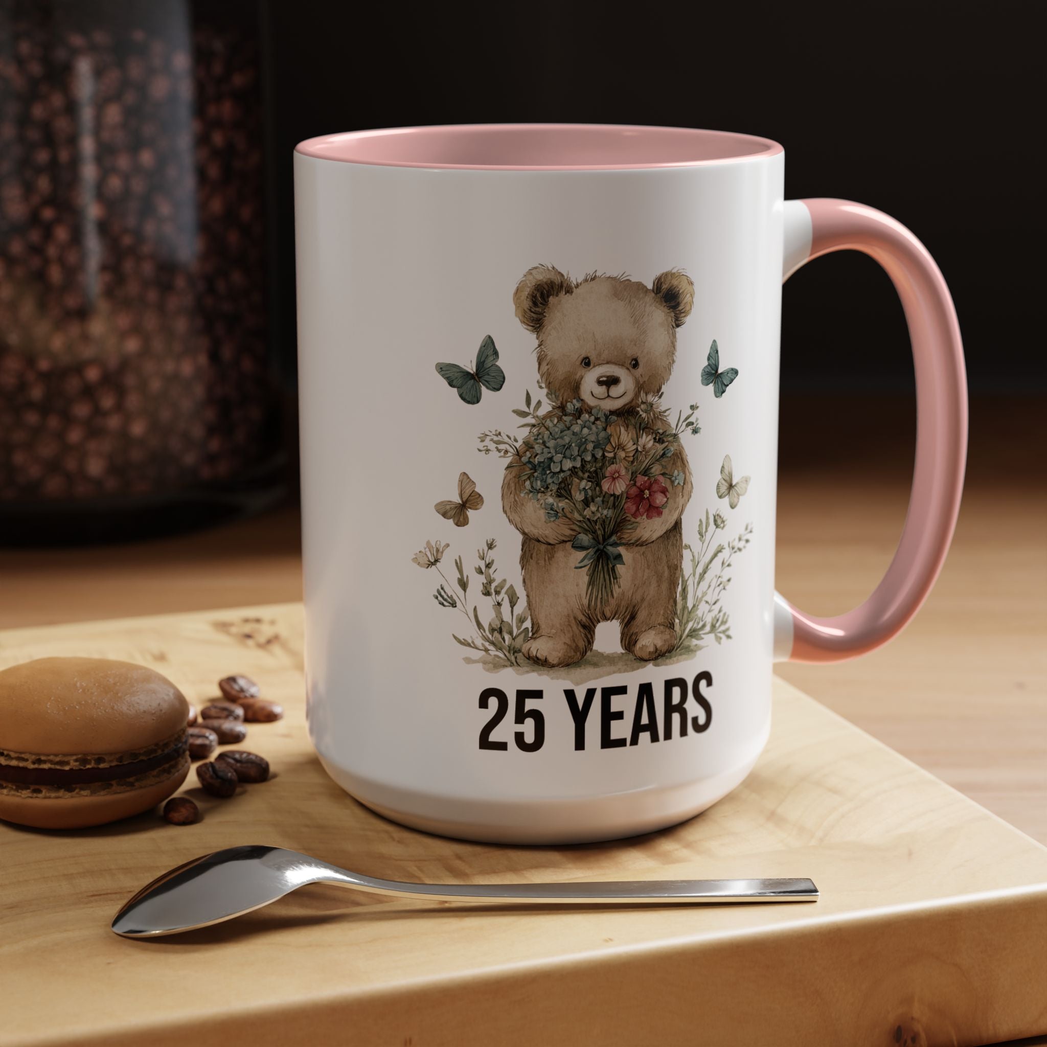 Mug with teddy bear design and '25 Years' text on a wooden surface with coffee beans and a macaron.