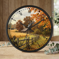 Round wall clock with a black frame and a scenic autumn landscape print featuring orange and yellow fall foliage, a rustic barn, and a wooden fence. The clock face has black Roman numerals and black hands, and it is styled on a wooden tabletop with eucalyptus leaves.
