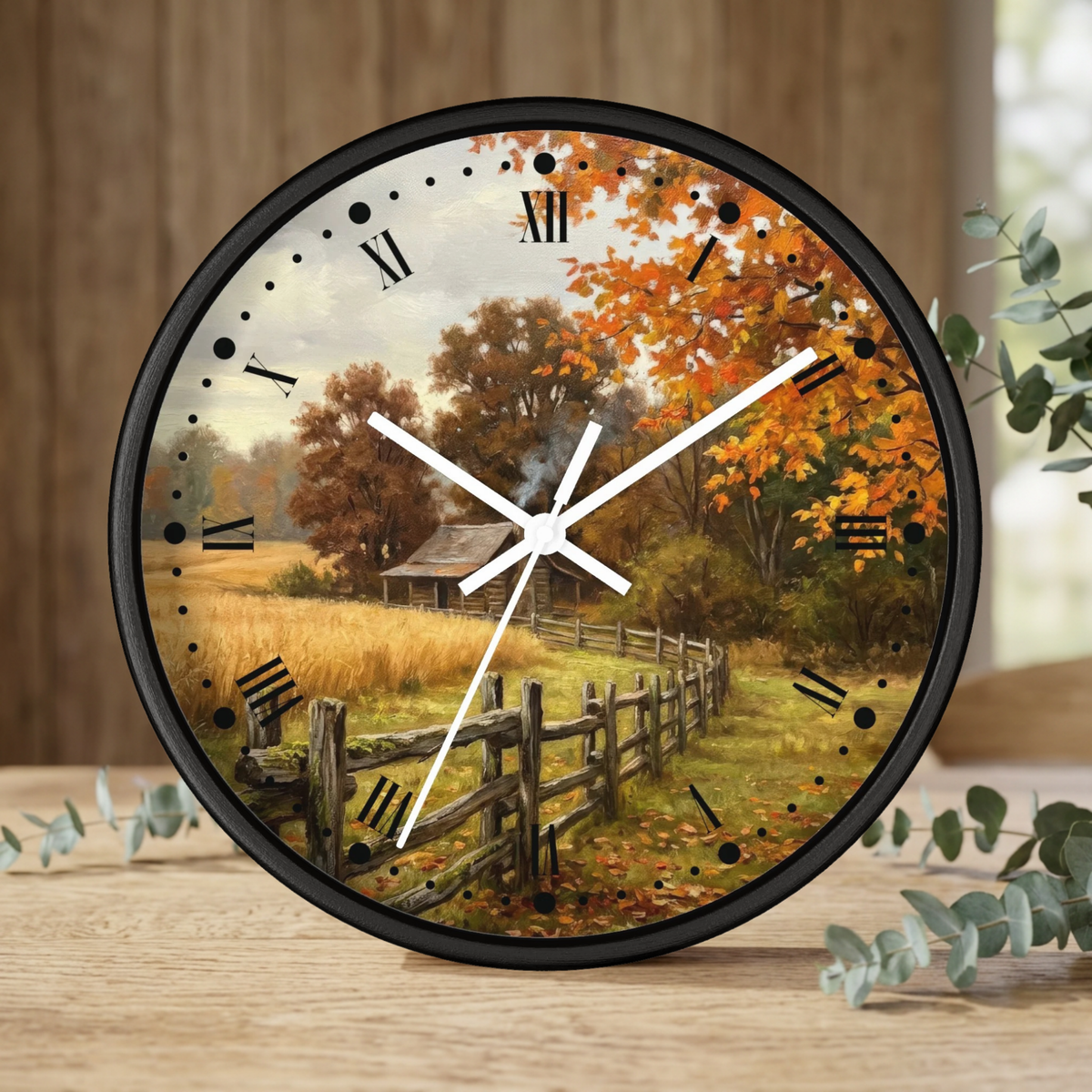 Round wall clock with a black frame and a scenic autumn landscape print on the face, featuring a wooden fence, trees with orange and yellow foliage, and a small cabin. The clock has black Roman numerals and white hands, shown standing on a wooden surface with decorative eucalyptus leaves in the foreground.
