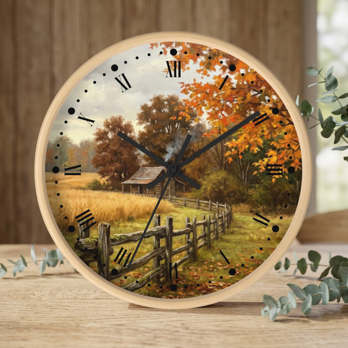 Round wall clock with a light wood frame and a scenic autumn landscape print featuring orange and yellow fall foliage, a rustic barn, and a wooden fence. The clock face has black Roman numerals and black hands, and it is styled on a wooden surface with eucalyptus leaves.