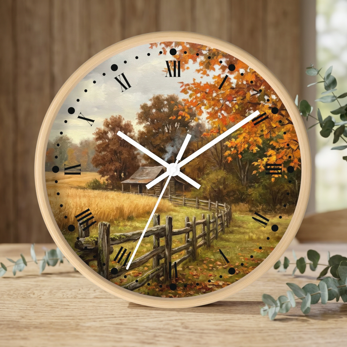 Round wooden clock with a scenic autumn landscape print, featuring a rustic fence, barn, and orange foliage, styled on a wooden tabletop with eucalyptus leaves.