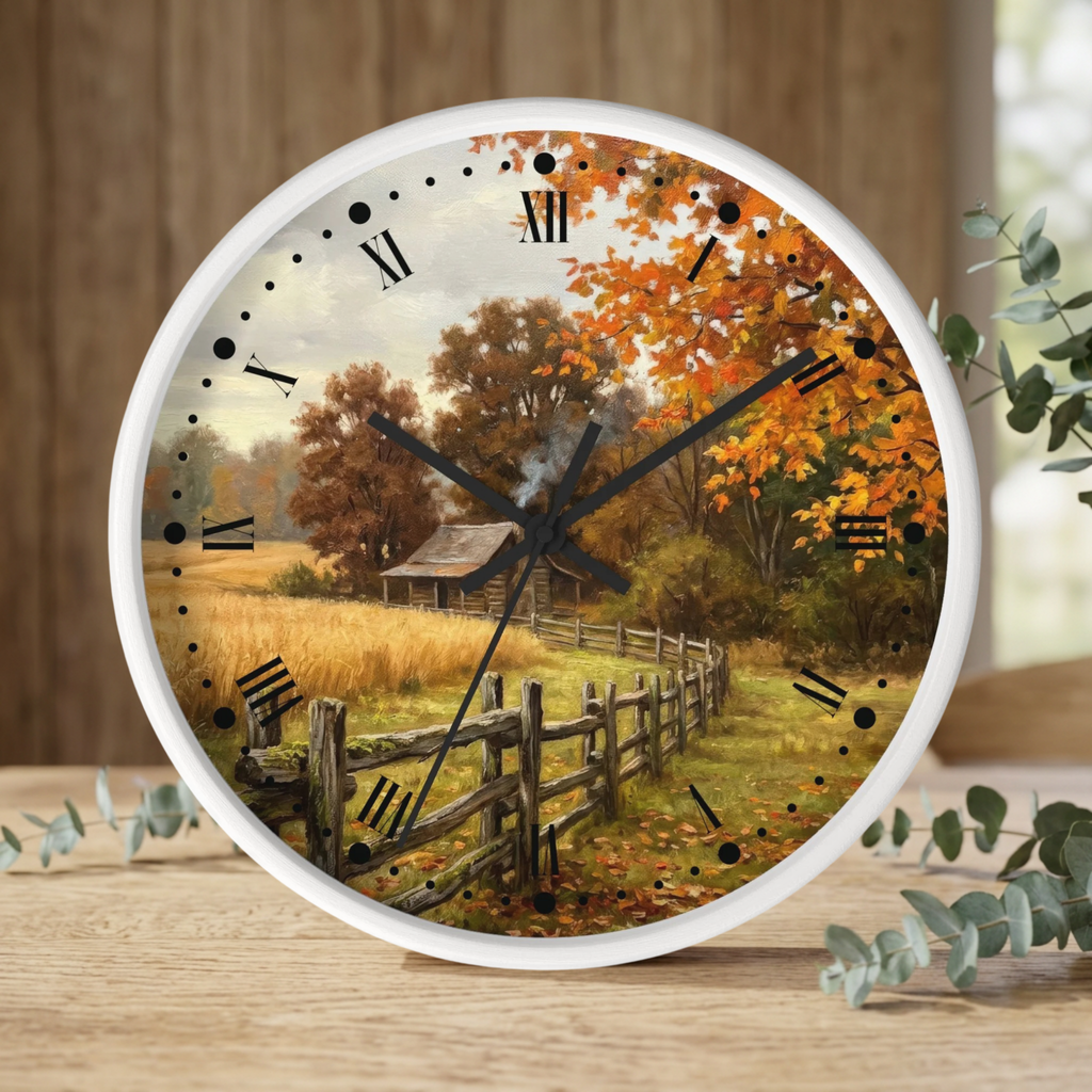 A round wall clock with a white frame and an autumn landscape print featuring orange and yellow fall foliage, a rustic barn, and a wooden fence. The clock is styled on a wooden tabletop with eucalyptus leaves and a blurred wood-paneled background.
