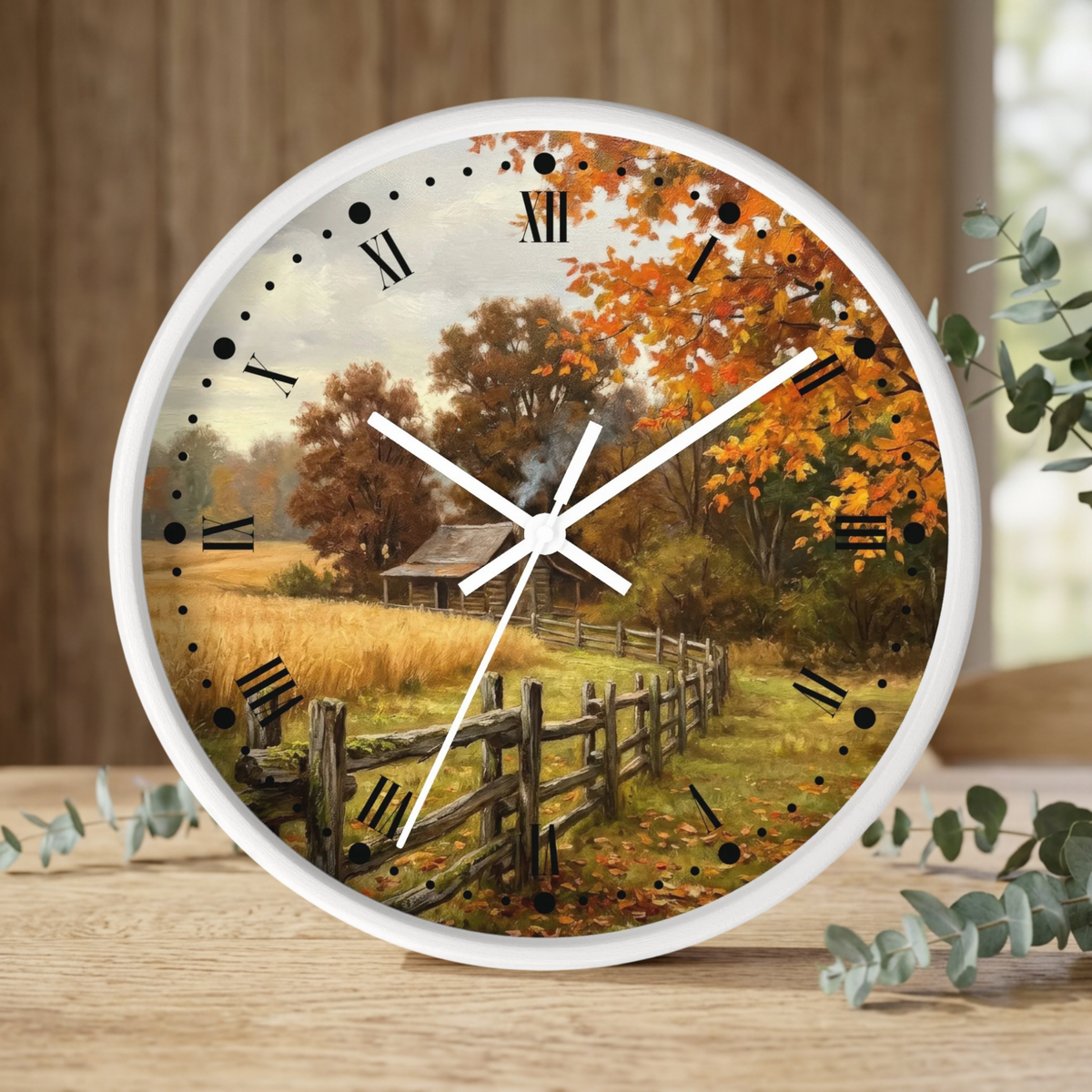 A round wall clock with a white frame and white hands, featuring a scenic autumn landscape print with orange trees, a wooden fence, and a small cabin. The clock is styled on a wooden tabletop with eucalyptus leaves and a blurred wood-paneled background.