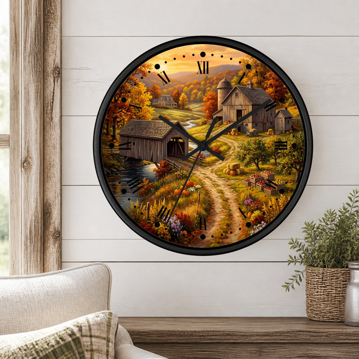 A round wall clock with a black frame and a detailed autumn farm landscape print, mounted on a white shiplap wall above a wooden table with a plant and part of a sofa visible.