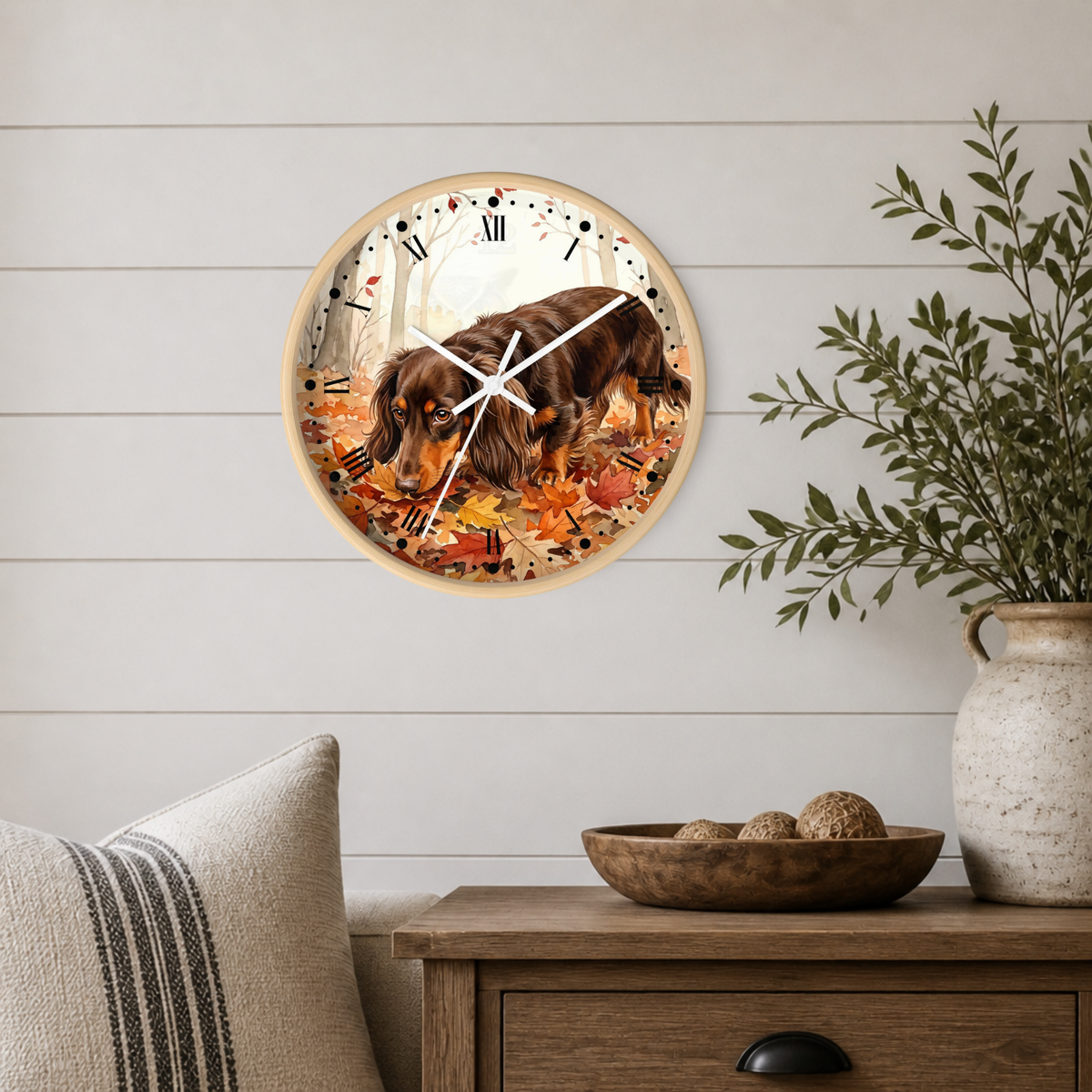 A round wall clock featuring a dachshund dog walking through autumn leaves, mounted on a white shiplap wall above a wooden side table with a vase and bowl.