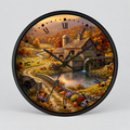 A round wall clock with a black frame and a detailed autumn landscape illustration on the face, showing a rustic mill, water wheel, stone bridge, flowers, and fall foliage. The clock has black Roman numerals and black hands on a light gray background.